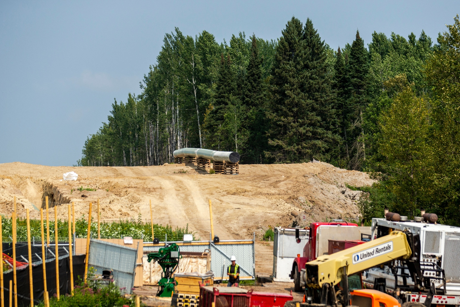 A stand of pine trees next to a bulldozed field where an unfinished pipe sits, next to construction equipment.