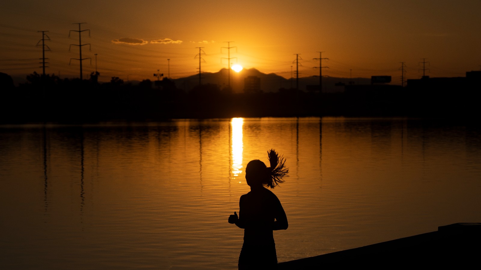 A woman runs along a lake as the sun rises next to power lines.
