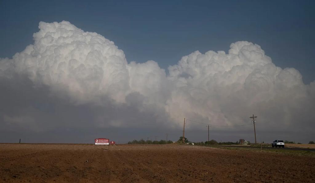Puffy clouds float above a brown field with a house in the distance.