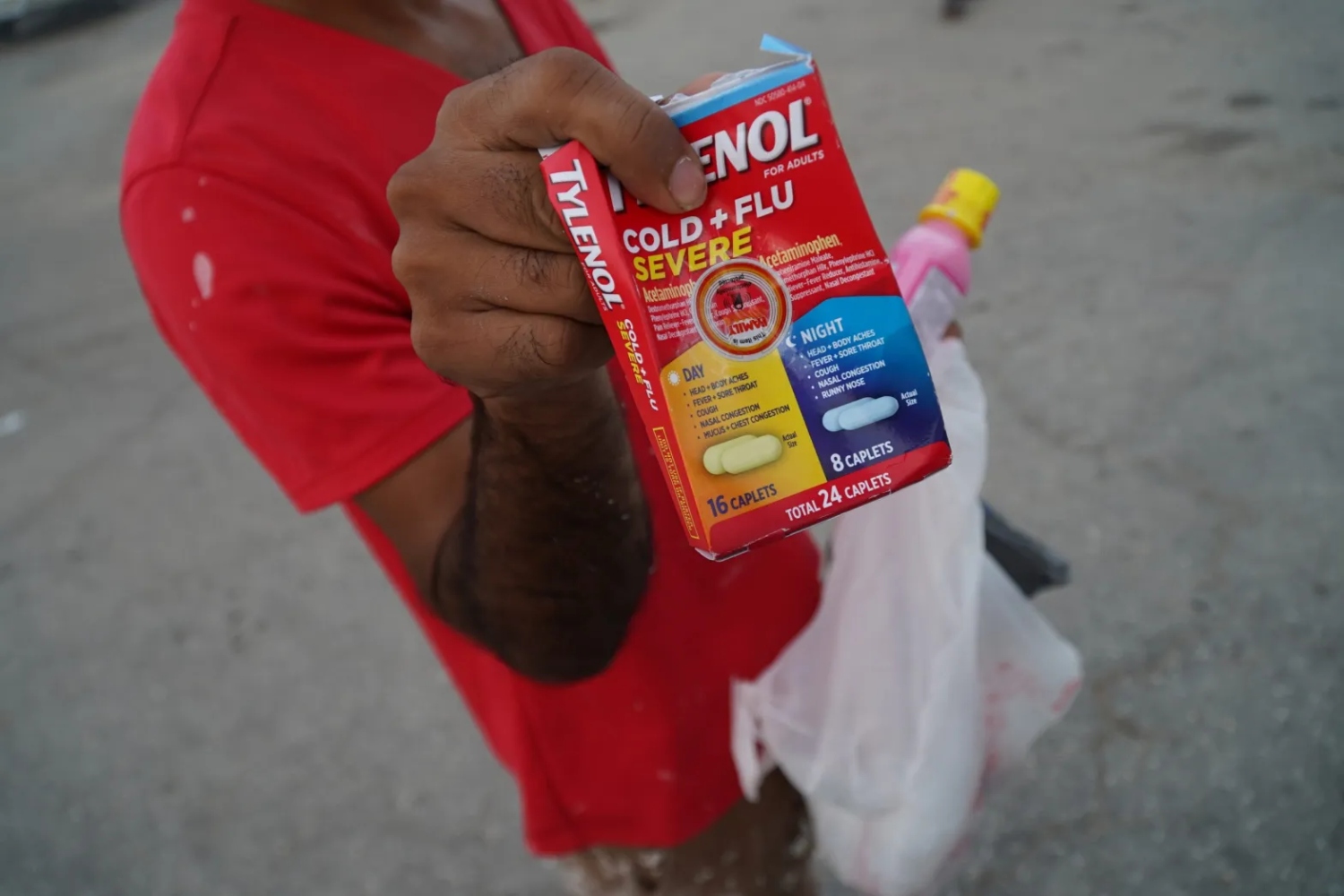 A restoration worker wearing a red shirt holds up a container of Tylenol Cold+Flu, with Pepto Bismol in his other hand. His face is not within the frame. The worker stands in a parking lot in Fort Myers where workers are picked up to clear out debris from structures destroyed by Hurricane Ian.