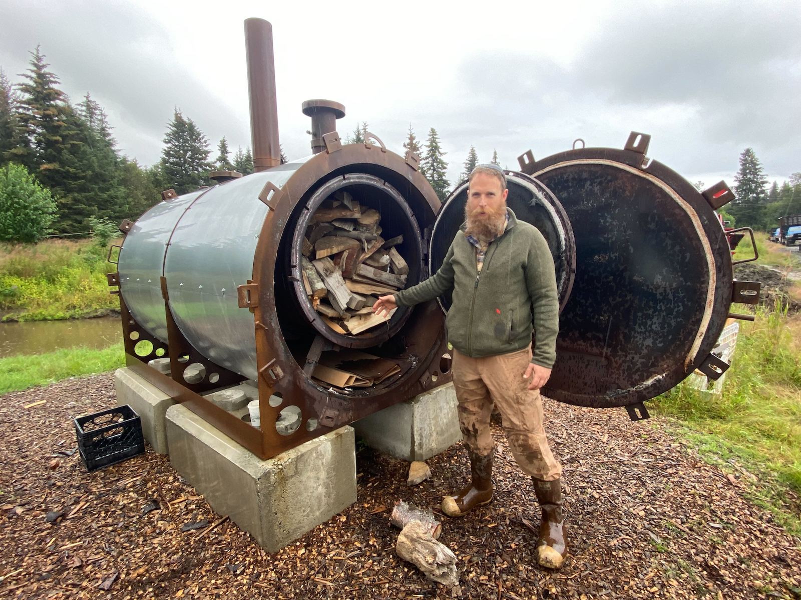A farmer stands in front of a biochar maker, stuffed with wood, at his farm in Homer, Alaska.