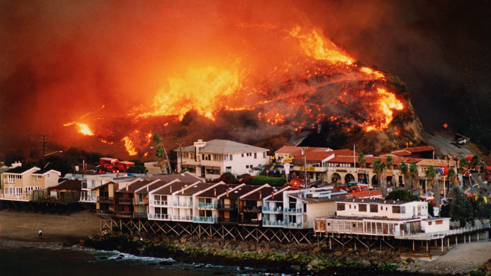 A wildfire burns on a wooded hillside behind a beachfront neighborhood in Malibu, California.
