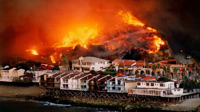 A wildfire burns on a wooded hillside behind a beachfront neighborhood in Malibu, California.