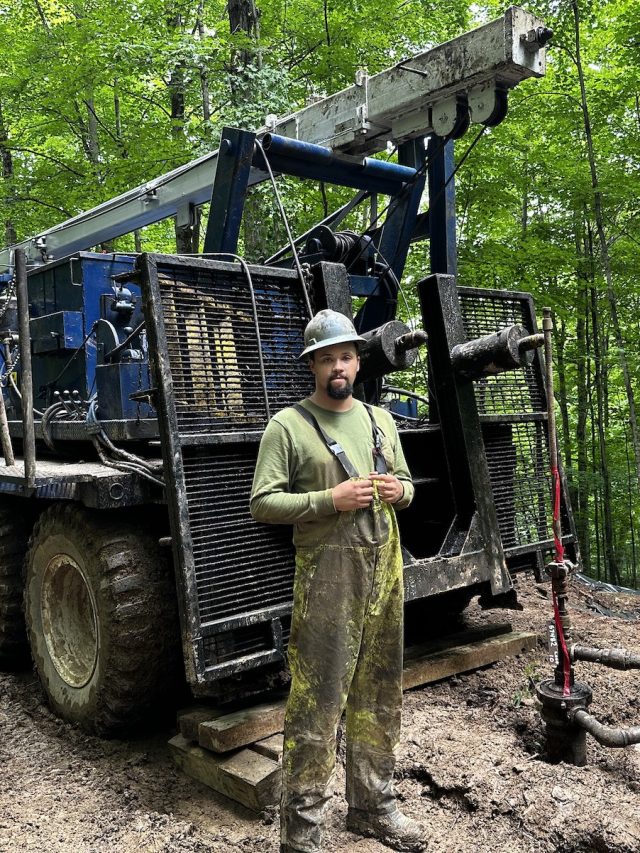 A man in overalls stands next to a large truck in the woods