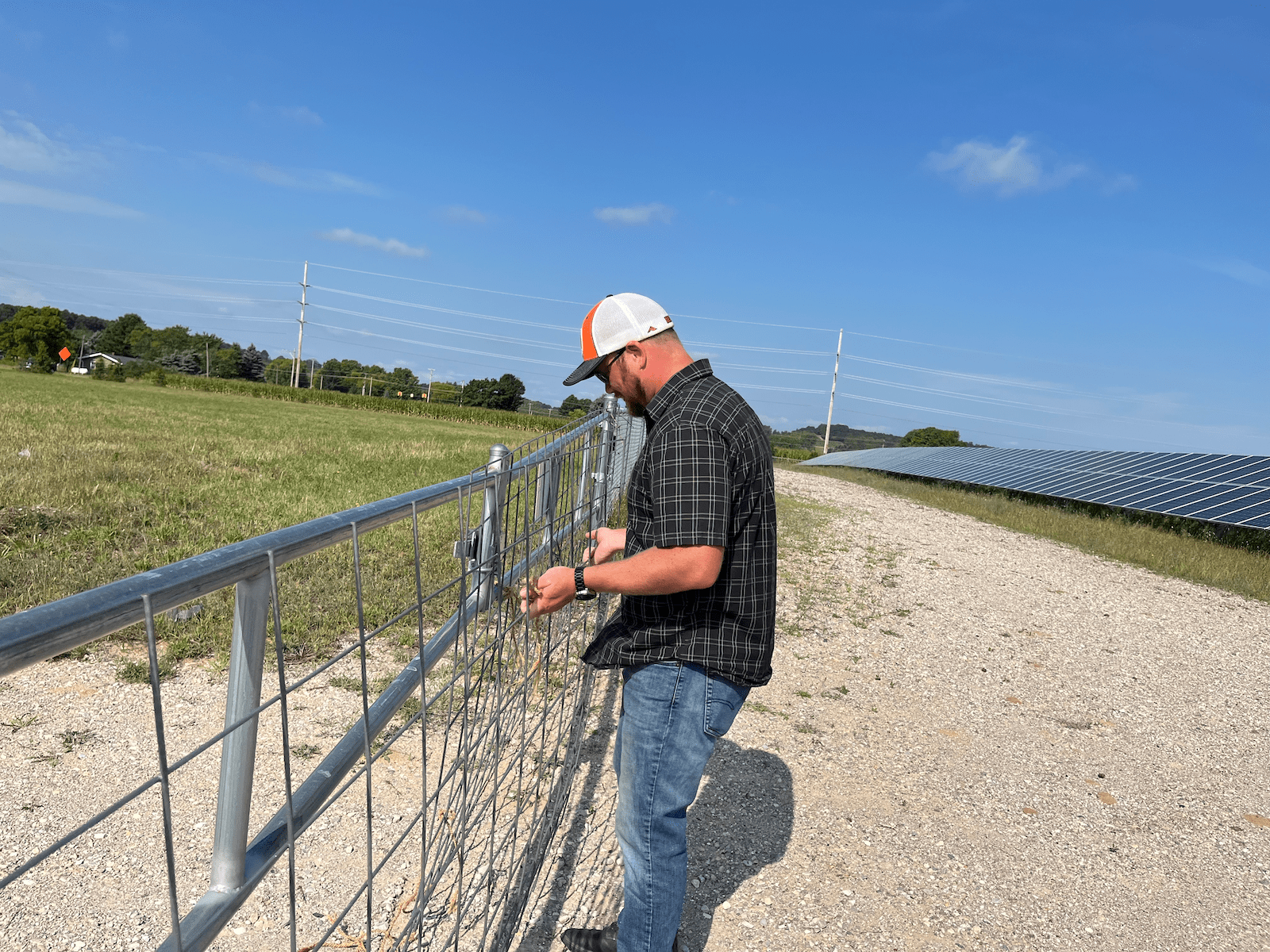Man in white trucker's cap and short-sleeve shirt stands on farmland and closes metal gate.