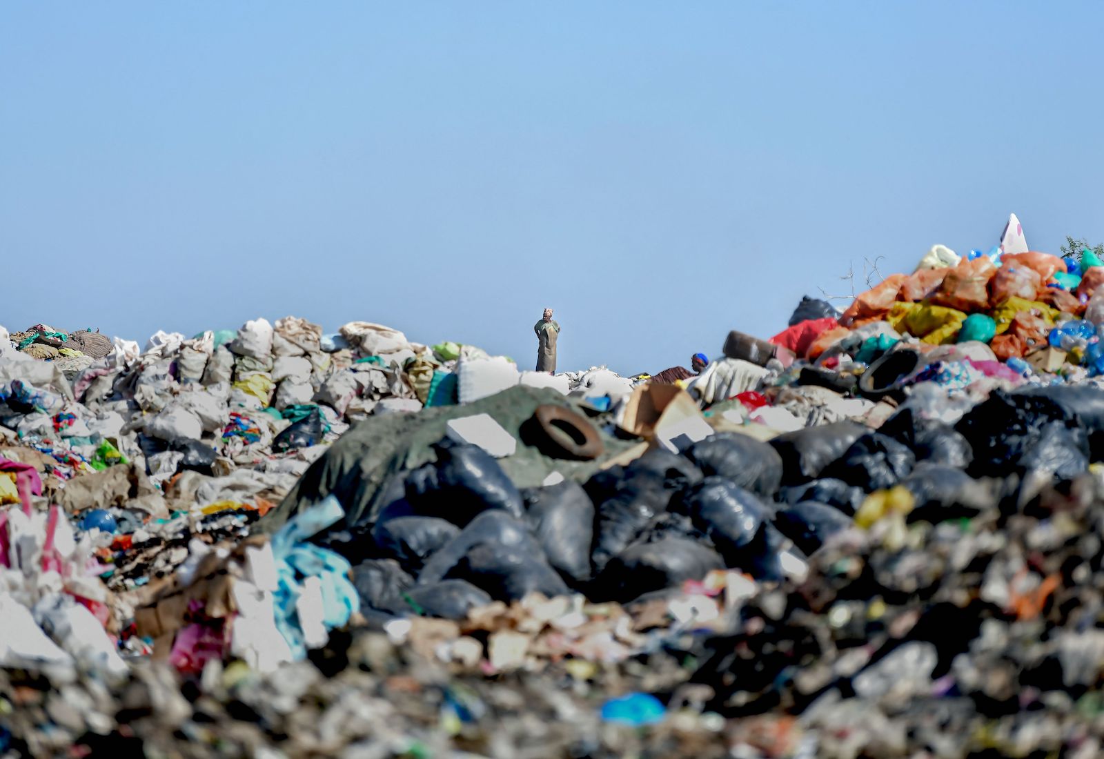 A waste picker stands in the distant background on top of a pile of trash