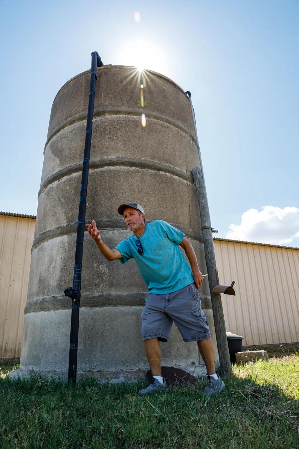A man in shorts, a blue tee shirt, and a baseball cap stands in front of a silo.