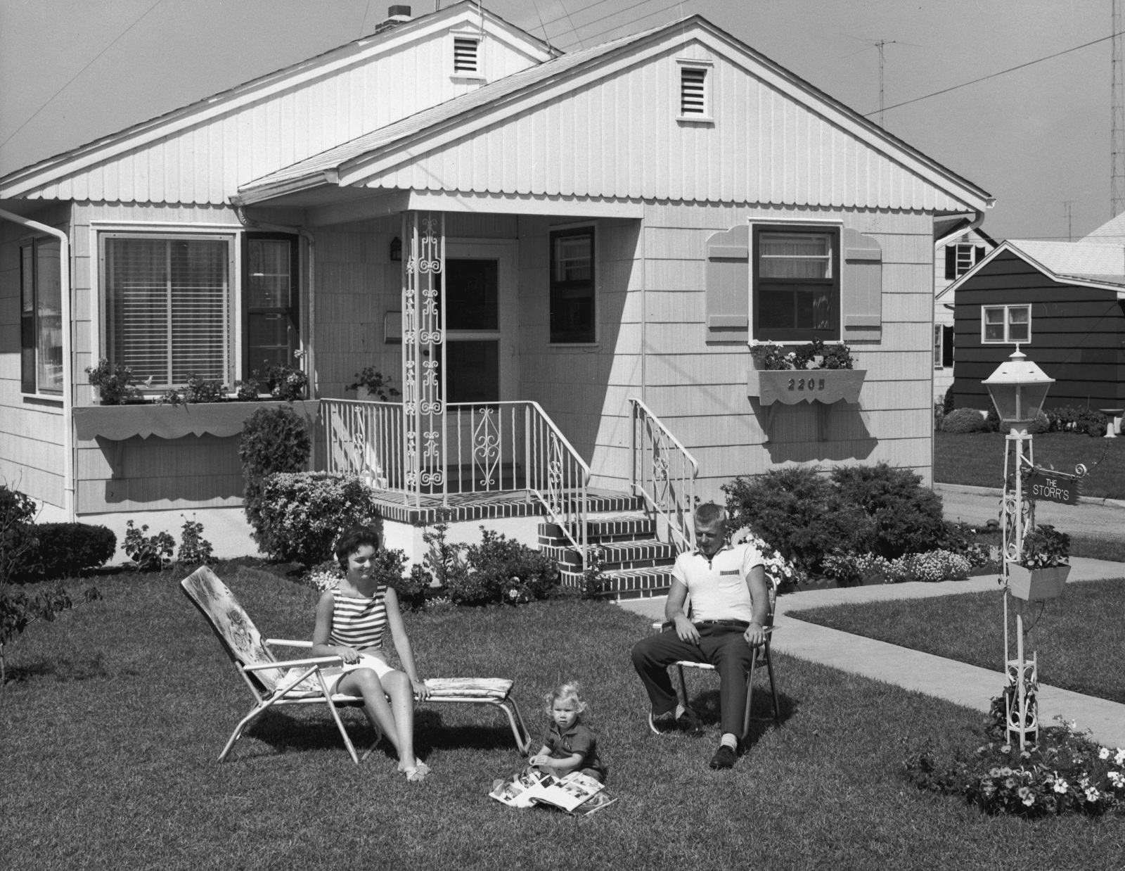 Black-and-white photo of a 1950s family sitting in lawn chairs in their yard.
