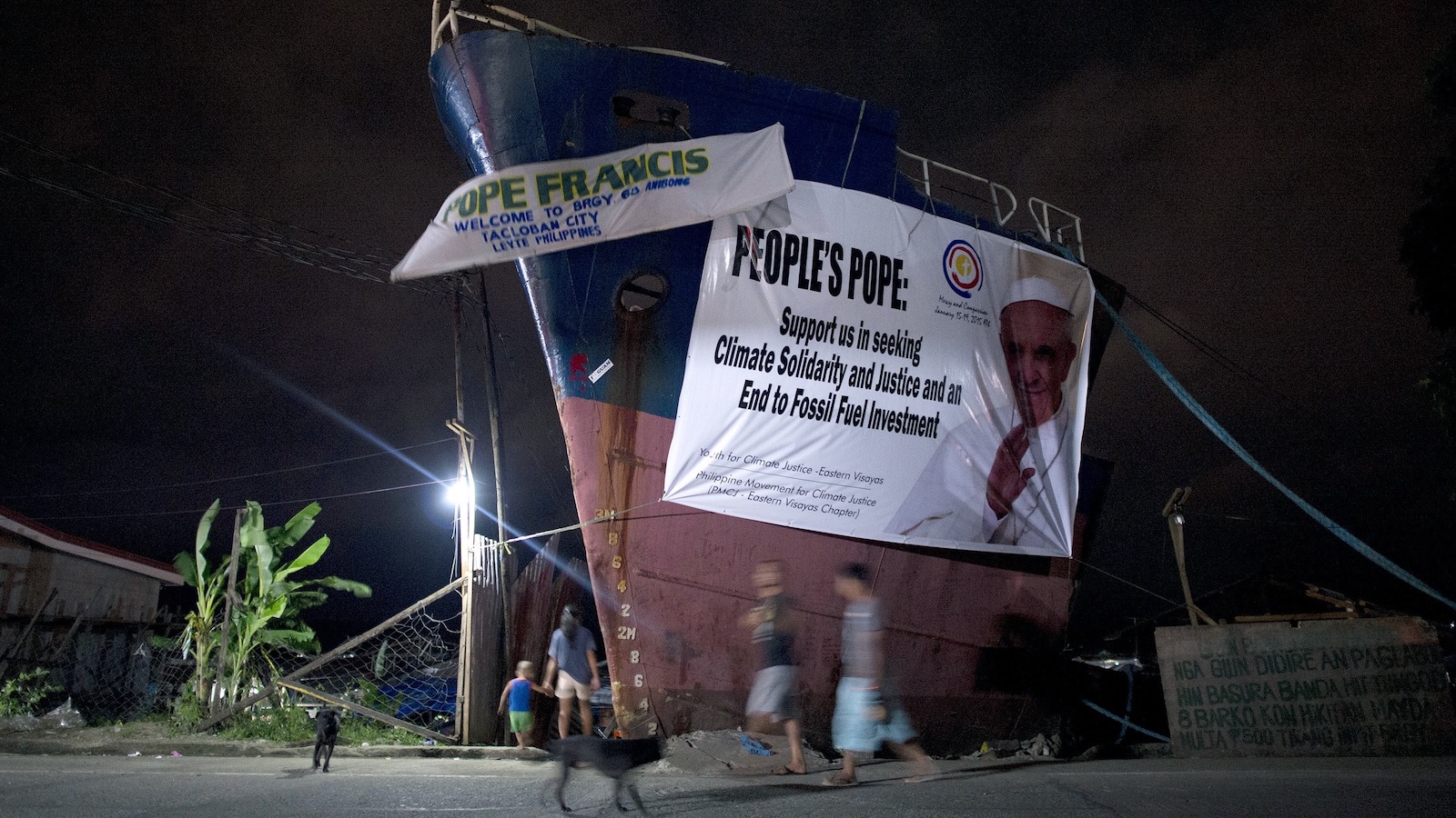 Residents of Tacloban in the Philippines walk past the bow of a broken ship that has washed ashore. The vessel bears a banner reading "People's pope: Support us in seeking climate solidarity and an end to fossil fuel investment."