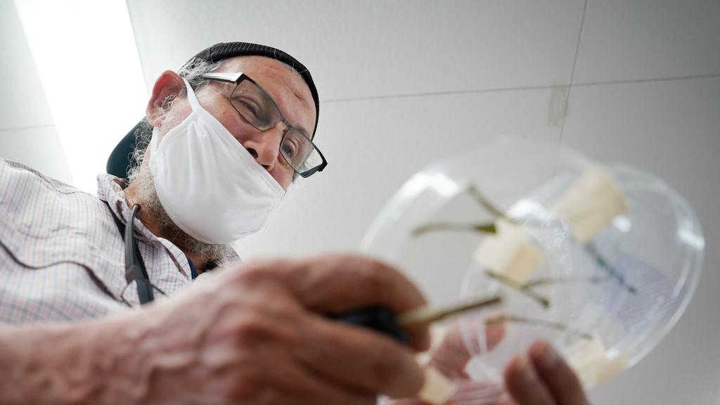 A man wearing a white face mask and a backwards baseball cap and glasses holds petri dishes.
