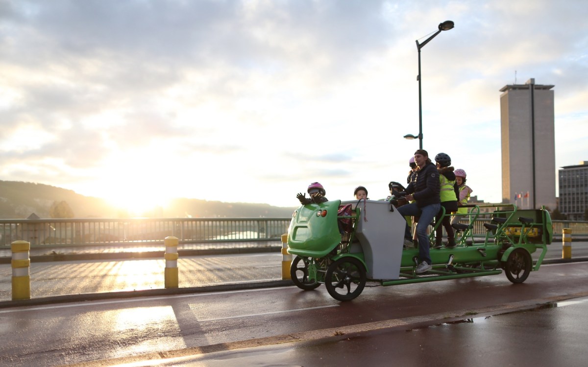 The sun streams over hills in the background of a bridge, where a group of small children and one adult ride on a four-wheeled green cart with several sets of pedals.