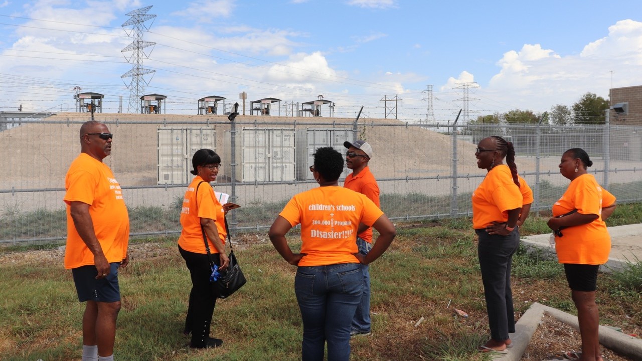 A group of Black women and men in orange shirts stand in front of an industrial facility.