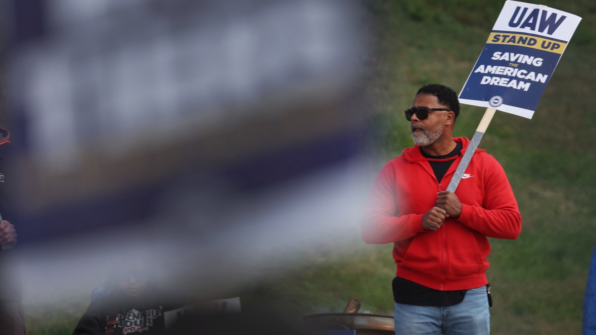 A worker holding a picket sign reading "UAW Stand Up. Saving the American Dream" walks a picket line outside a Ford factory.