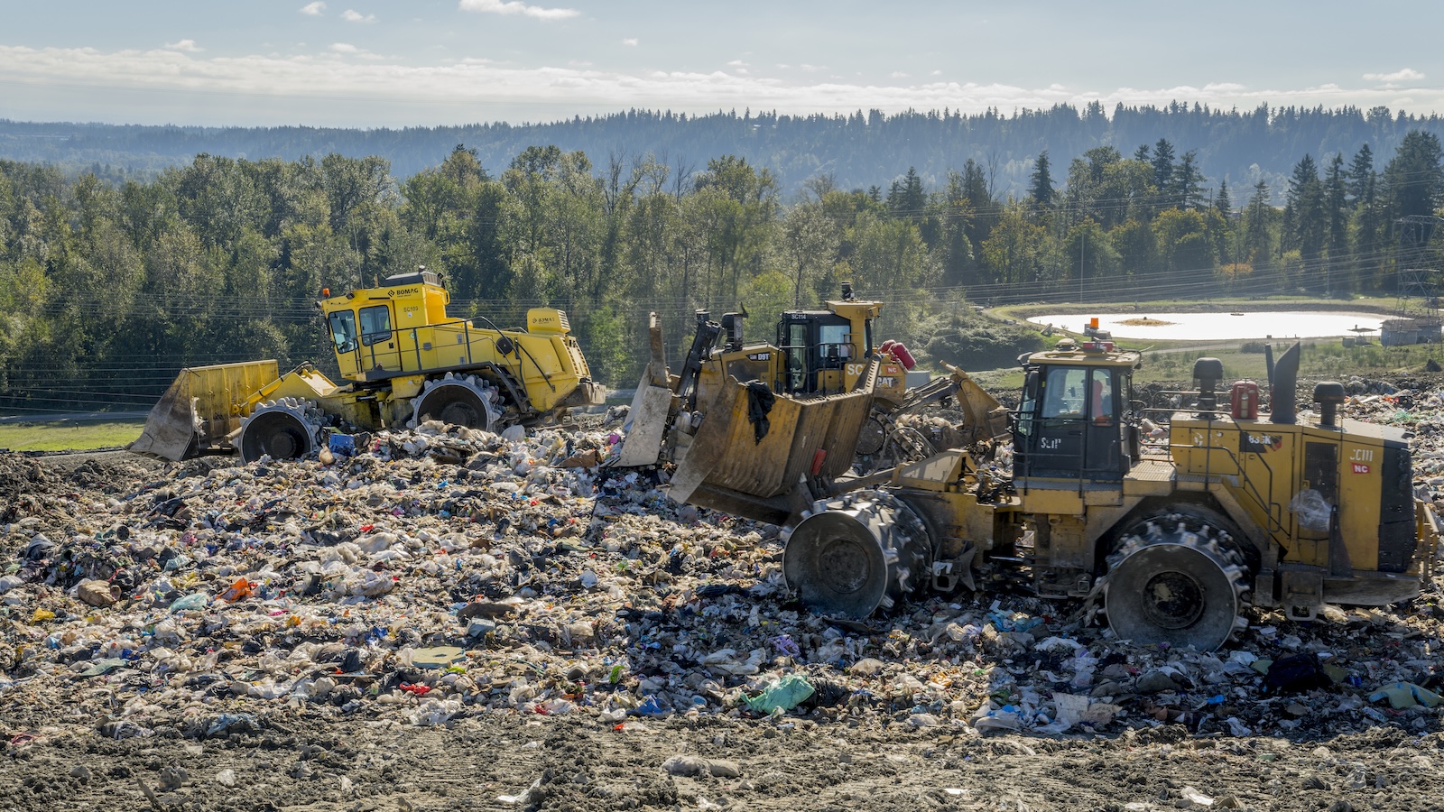 Tractors sit on top of a landfill, with a forest in the background