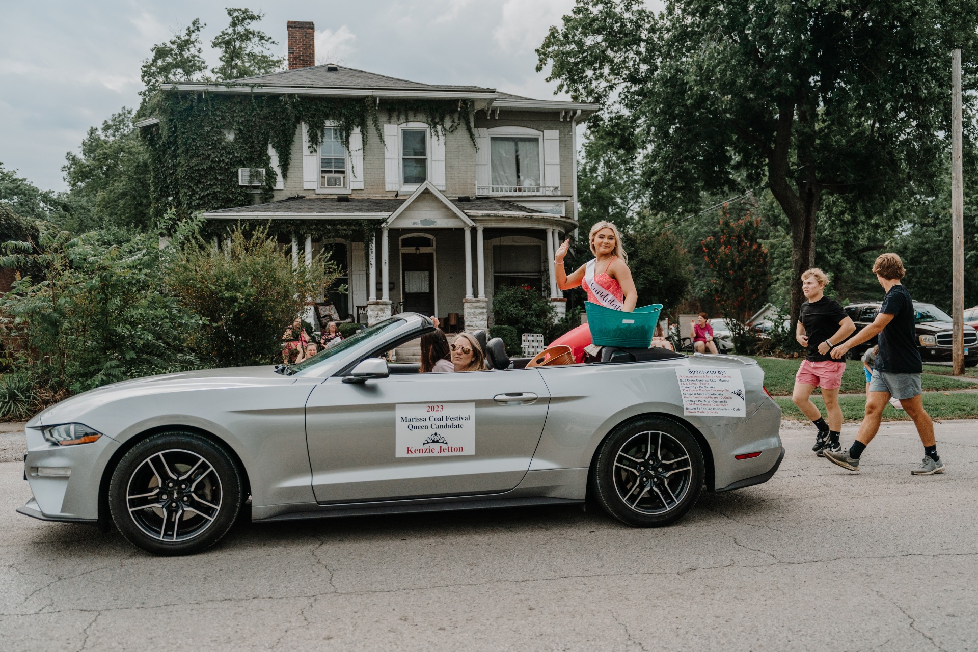 A young woman in a pink dress rides in the back of a grey convertible