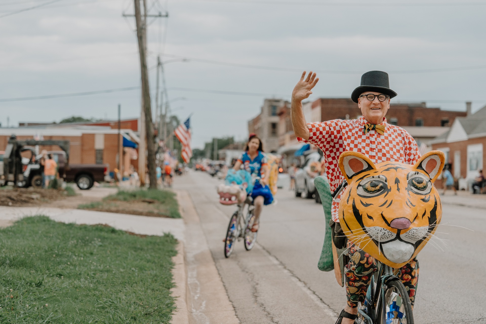 A man in a top hat waves at the camera while riding a bike with a paper mache tiger head attached to the front