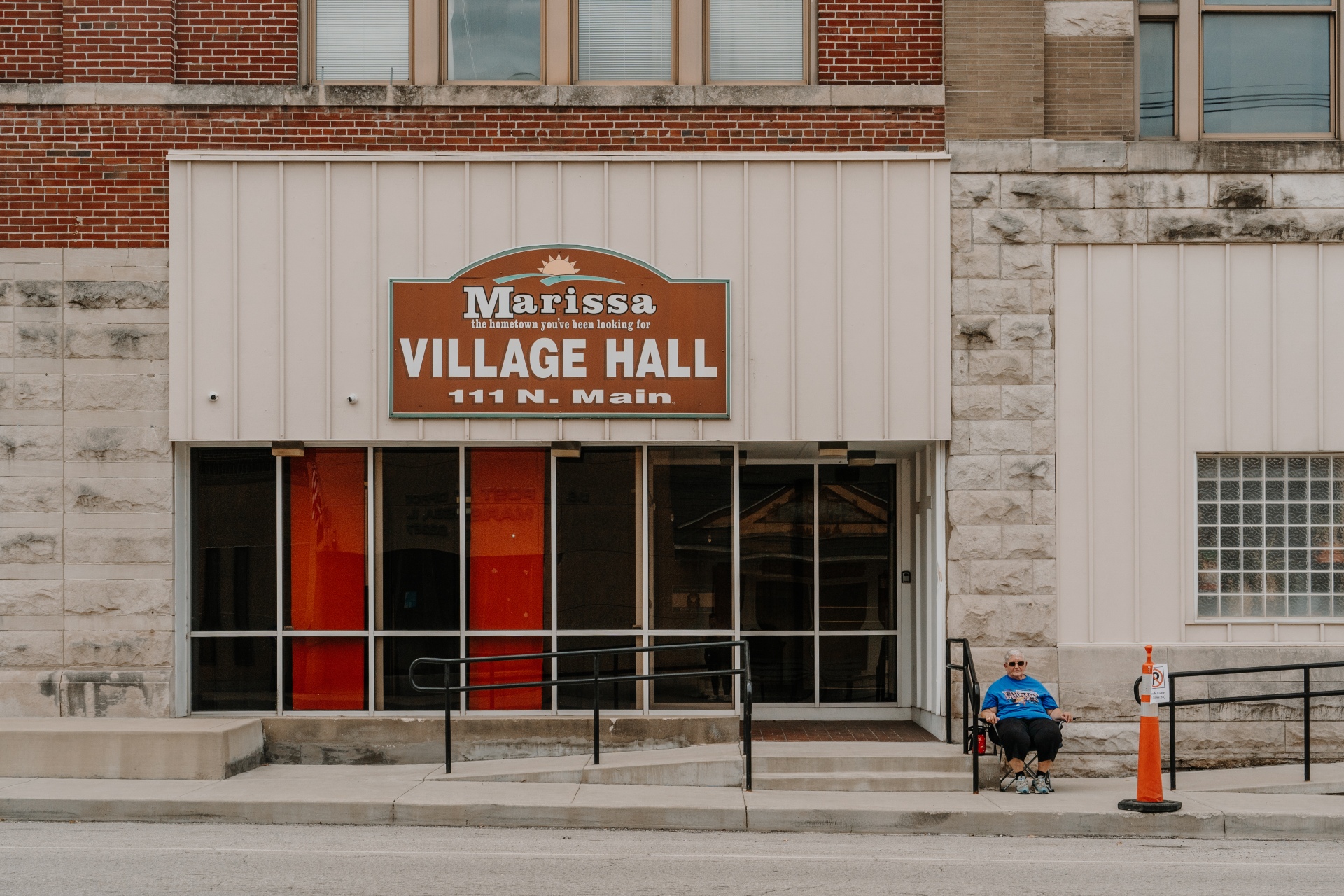 Rosemary Fulton sits in a folding chair next to the doorway of a large stone and brick building. A brown sign above the doorway reads: Marissa, the hometown you've been looking for. Village Hall. 111 N. Main.
