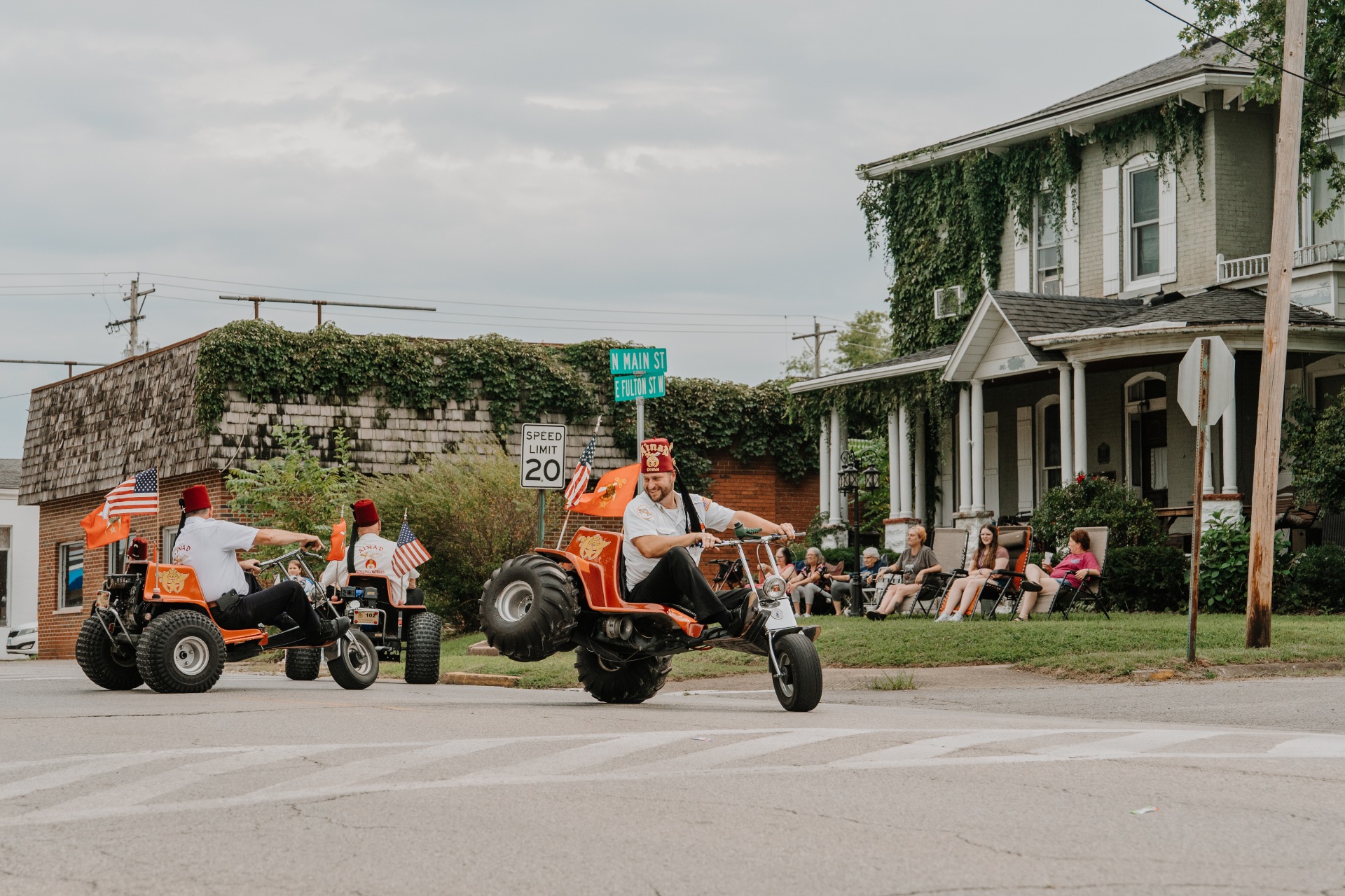 Men ride ATV bikes down a residential street as people watch from the lawn