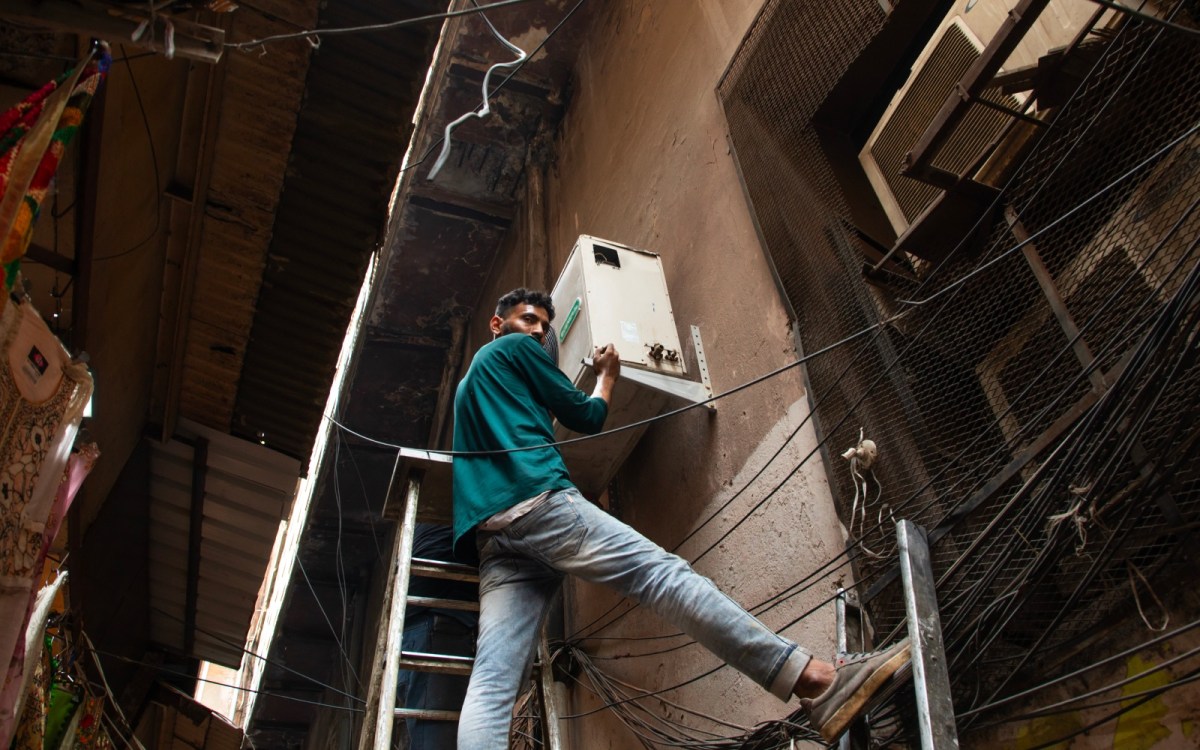 A man stands with his feet on two different ladders, installing an AC on the side of a stone building.