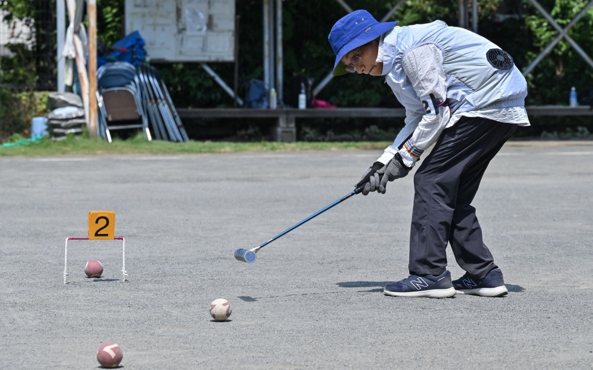 An older person wearing a blue bucket hat and a puffy jacket with visible fans crouches over a set of balls, holding a mallet.