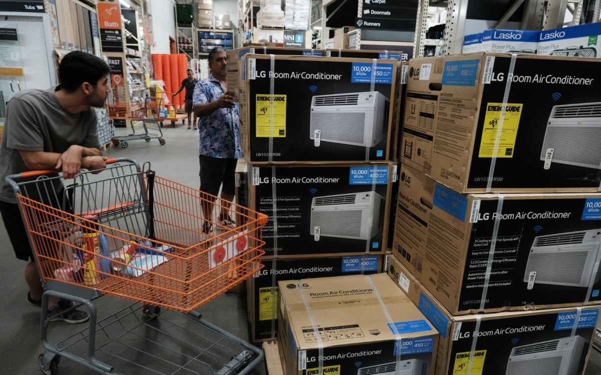 A man stands leaning over an orange grocery cart while another lifts a boxed AC from a stack.