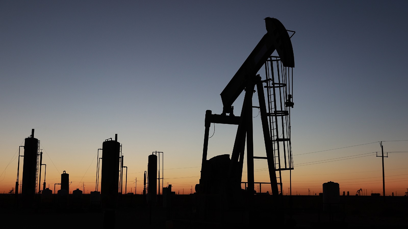An oil pumpjack and industrial towers are silhouetted against a setting sun.