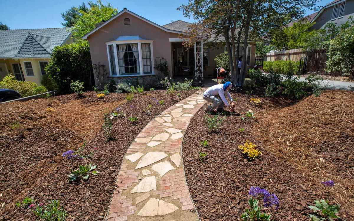 A stone pathway in the foreground leads to a small house; a man is crouched in the mulch yard with his hands in the ground. A few plants are scattered throughout.