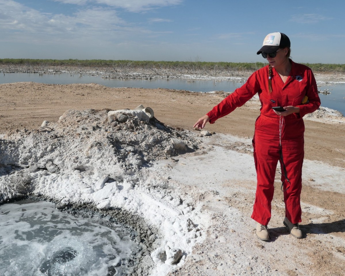 A woman in a baseball cap and red coveralls points to a large puddle.