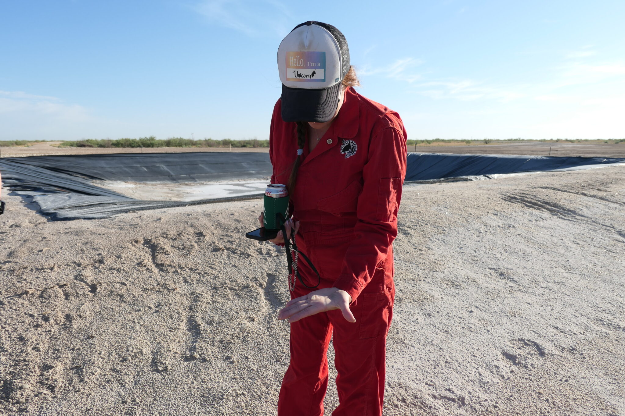 A woman in a red jumpsuit and baseball cap stands in sand looking at something in her hand.