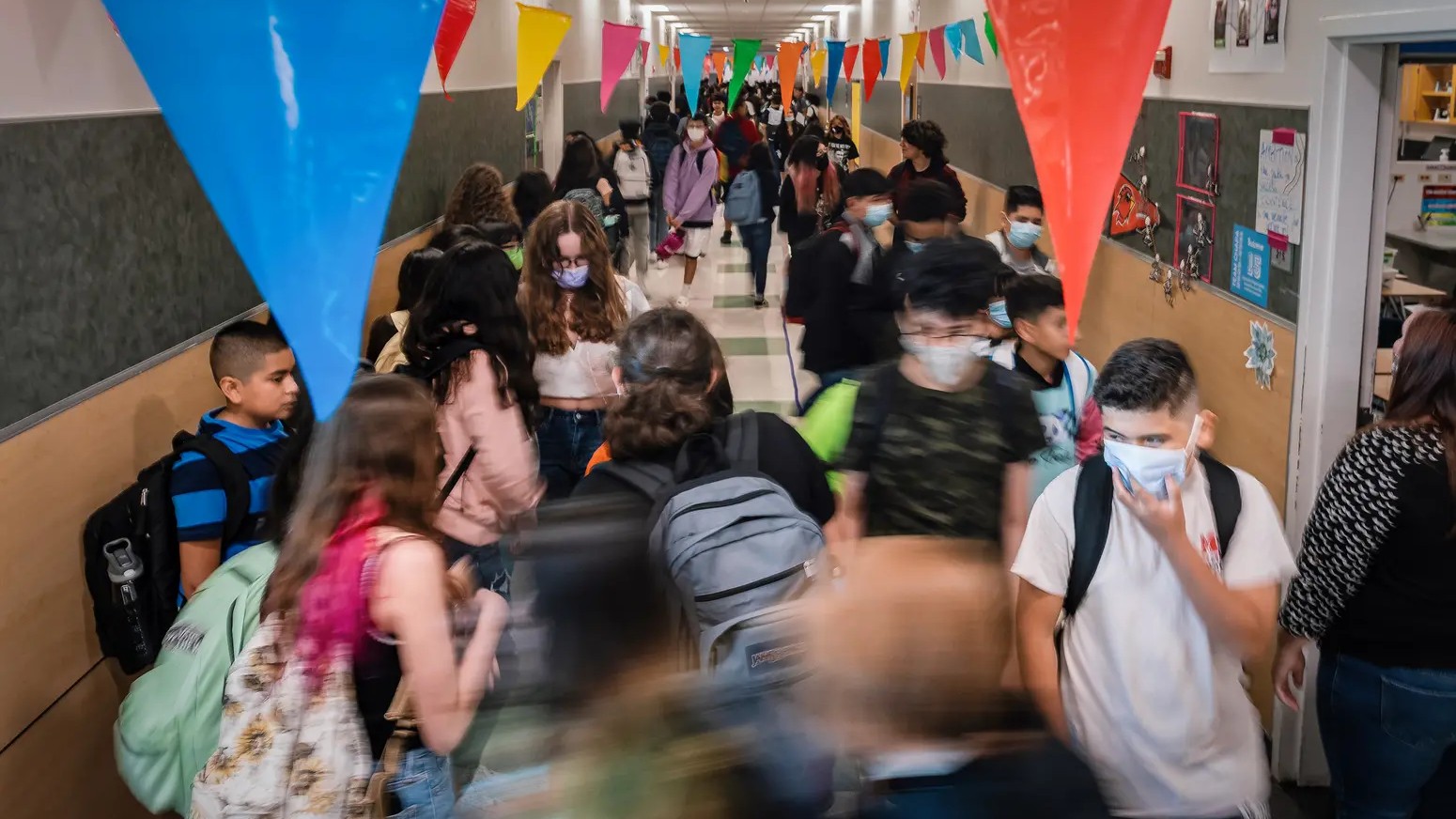 A busy hallway bedecked with flags is filled with students wearing backpacks and masks.