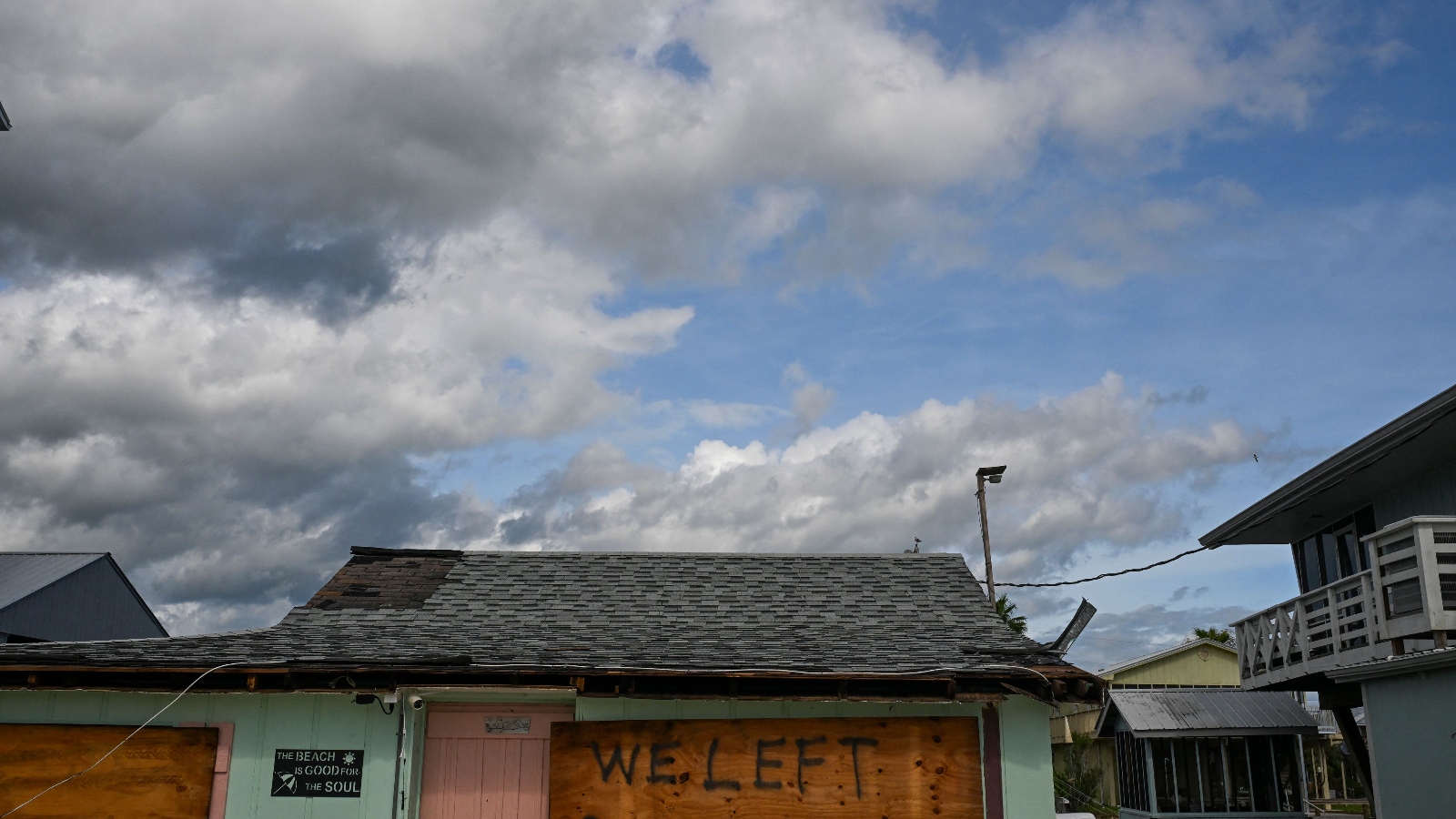 A pastel green house is boarded up after Hurricane Idalia made landfall in Florida. The board reads WE LEFT under a slightly damaged roof. The sky is blue with large, white clouds.