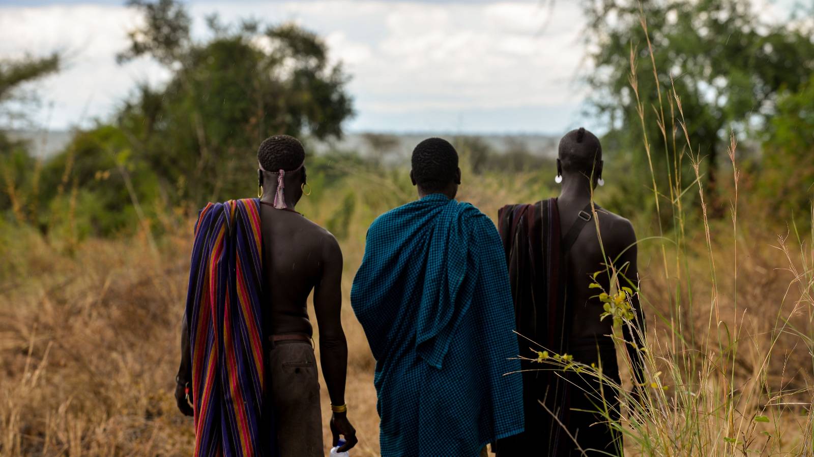Three members of the Mursi tribe in Ethiopia look out at a forest in front of them, their backs to the camera.