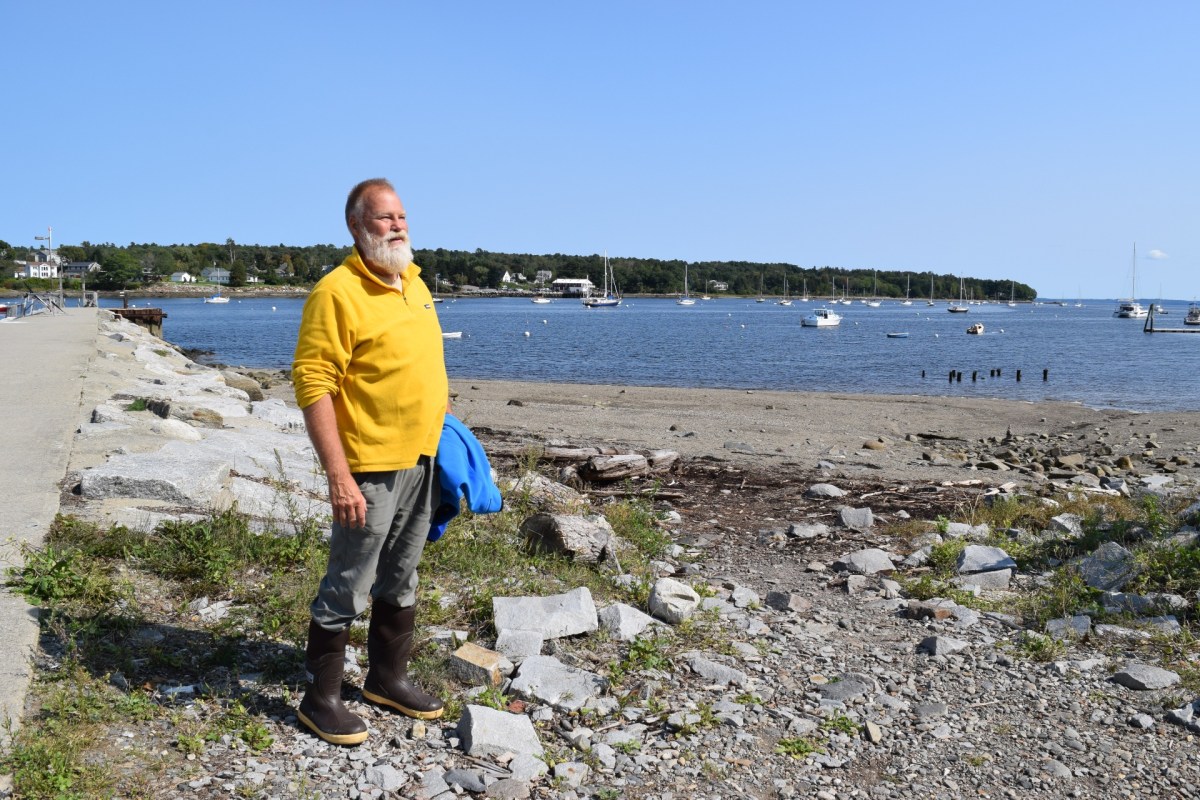 A man with a grey beard, yellow shirt, jeans, and rubber boots, stands on a rocky beach
