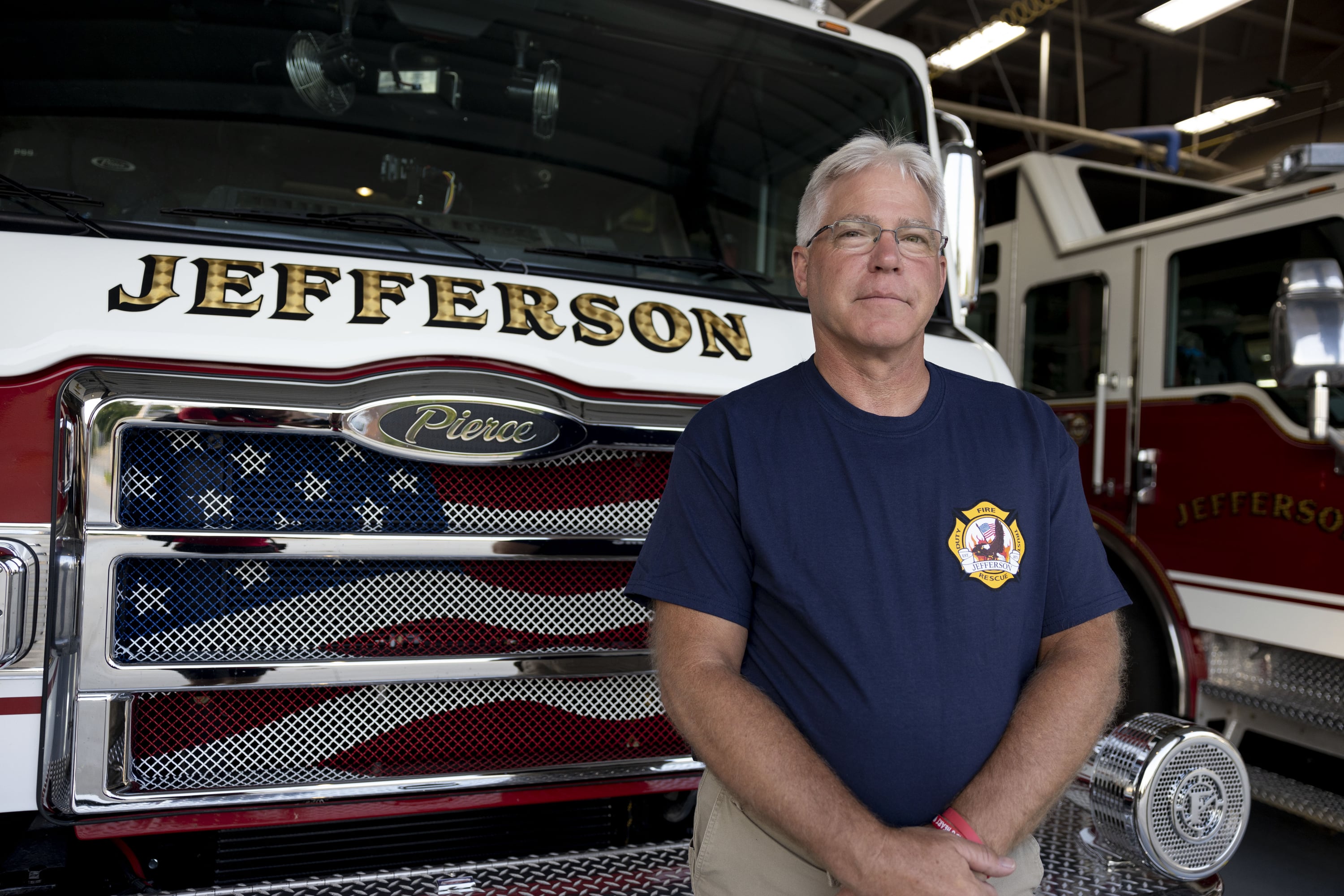 An older man in a blue shirt and glasses stands in front of a firetruck.