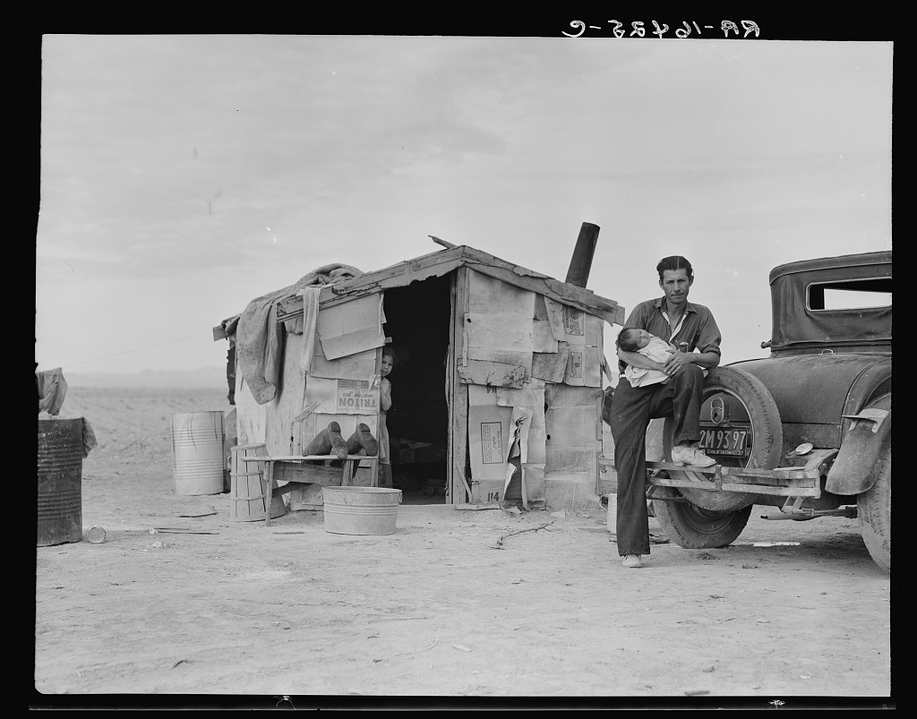 A man holds a baby outside of a shack and stands with his foot on an old-fashioned car.