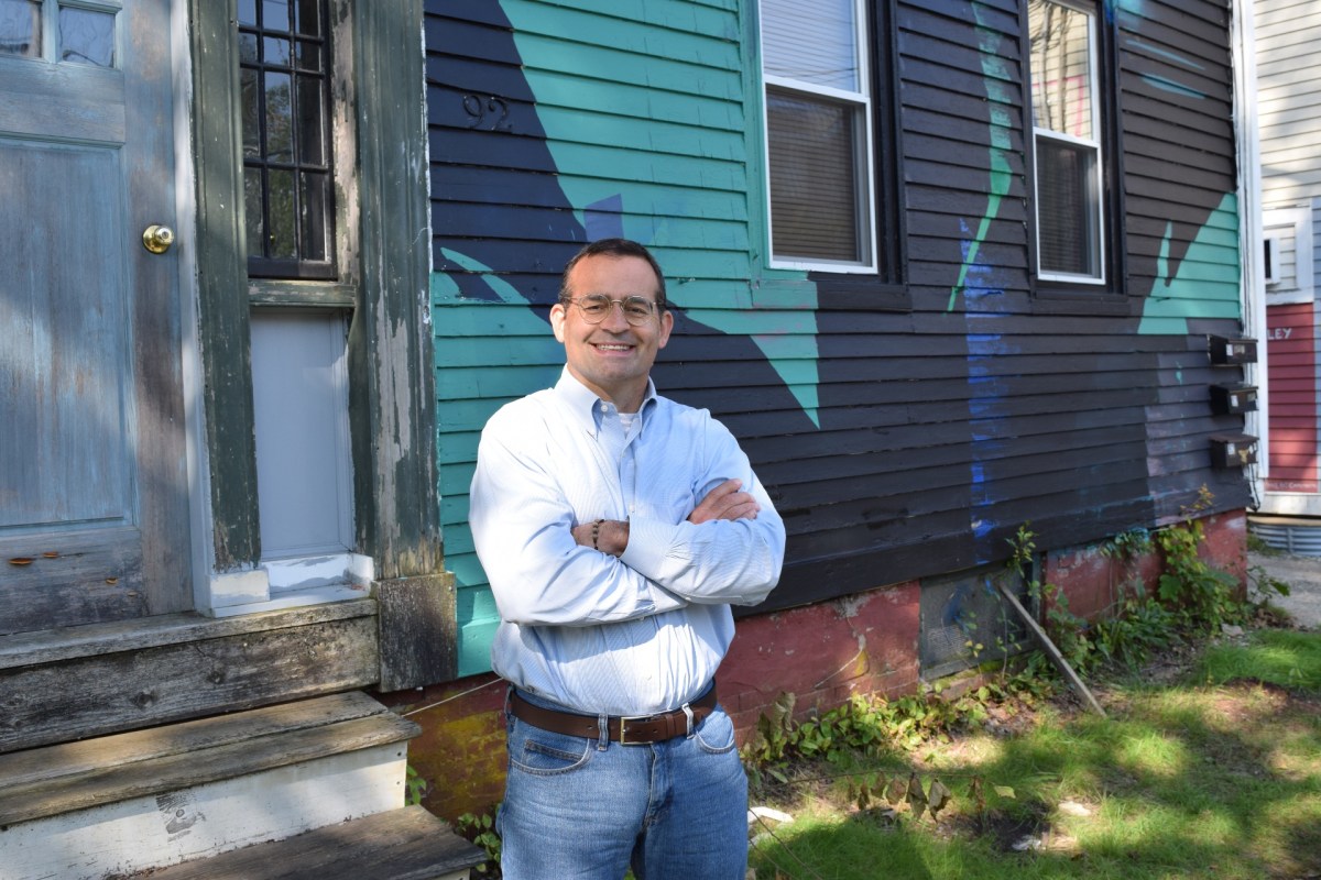 A man in a light blue shirt tucked into jeans smiles with his arms folded, standing in front of a wooden building painted in bright colors