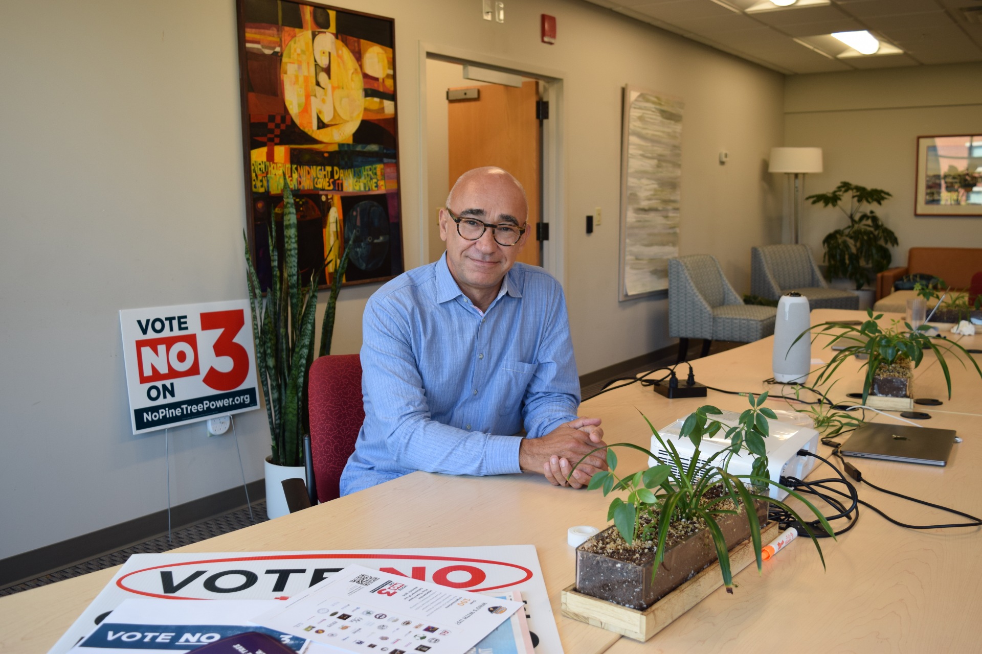 A man in a blue collared shirt and glasses sits at a table in an office. Posters around him read 'Vote No on 3'