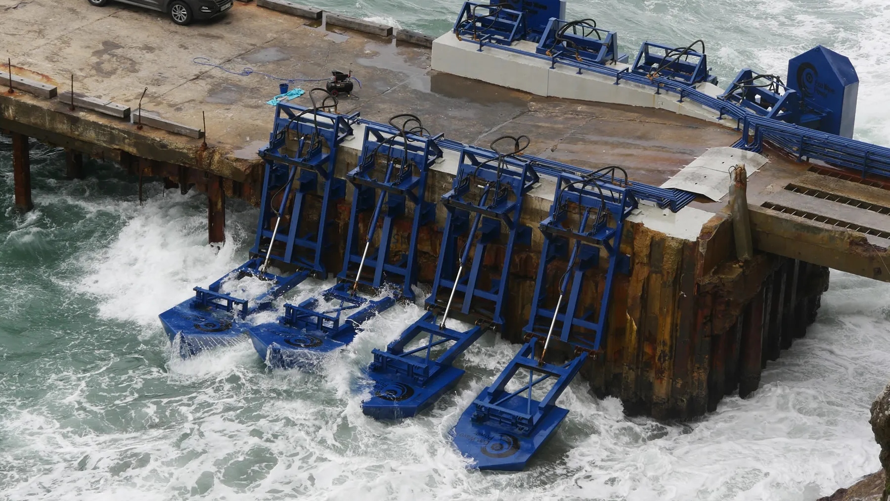 Four blue metal contraptions attached to a pier are battered by waves.