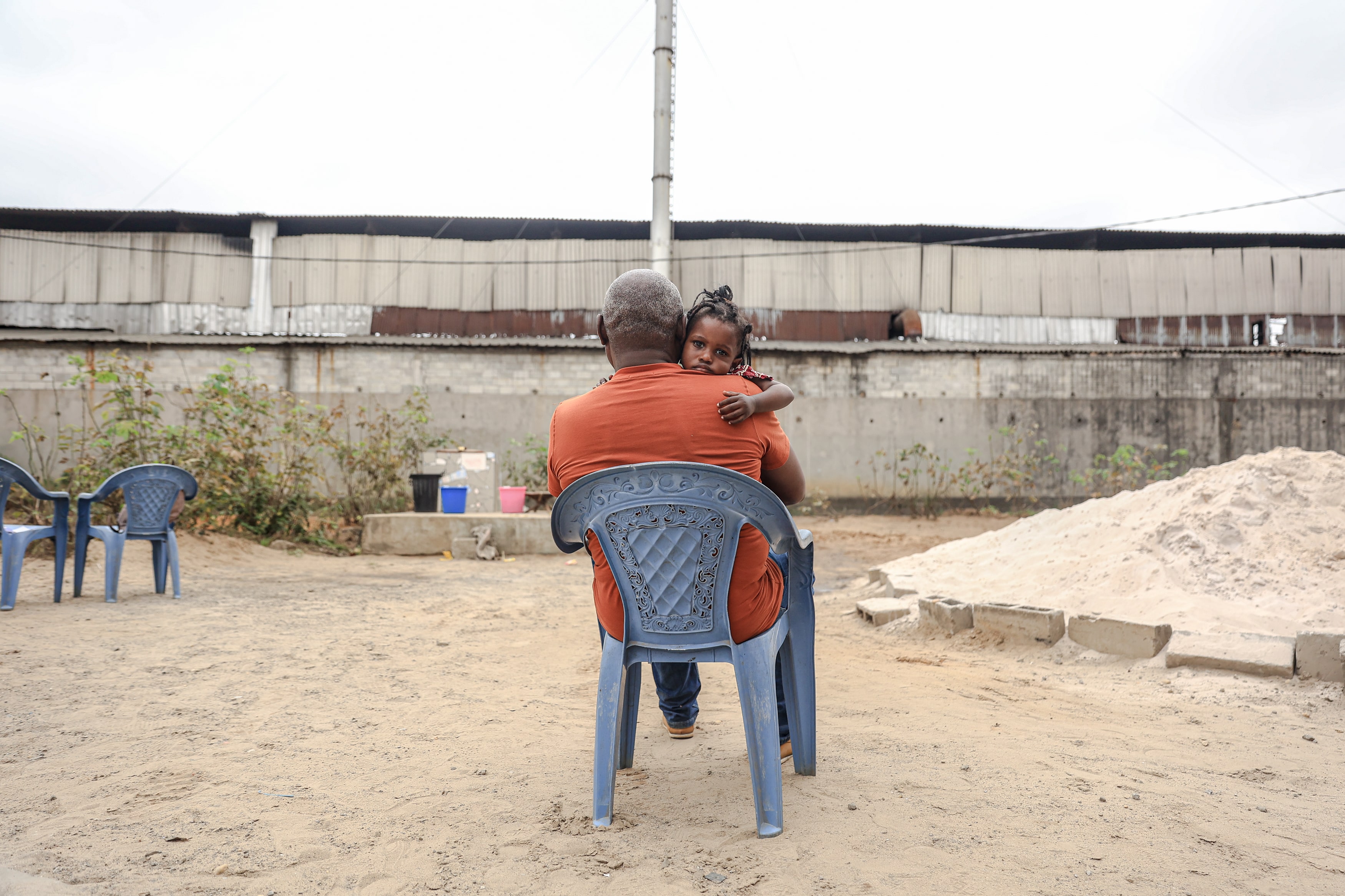 A little girl hugs a man who's sitting on a plastic chair in a sandy yard.