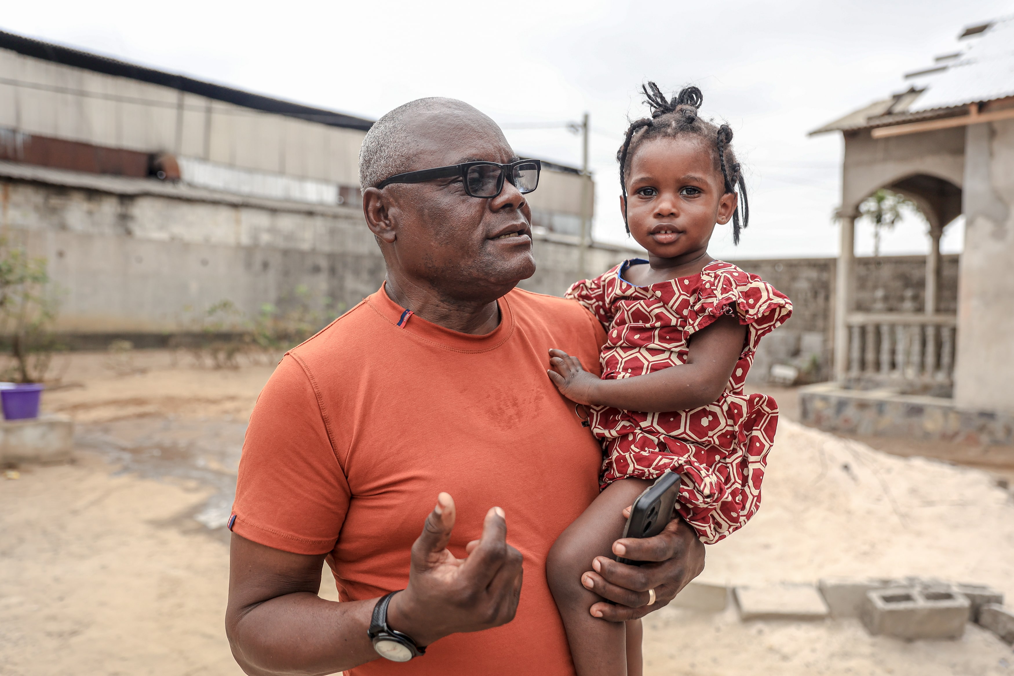 A Black man in an orange tee shirt and black glasses holds a little girl with braids and a red dress in his arms.