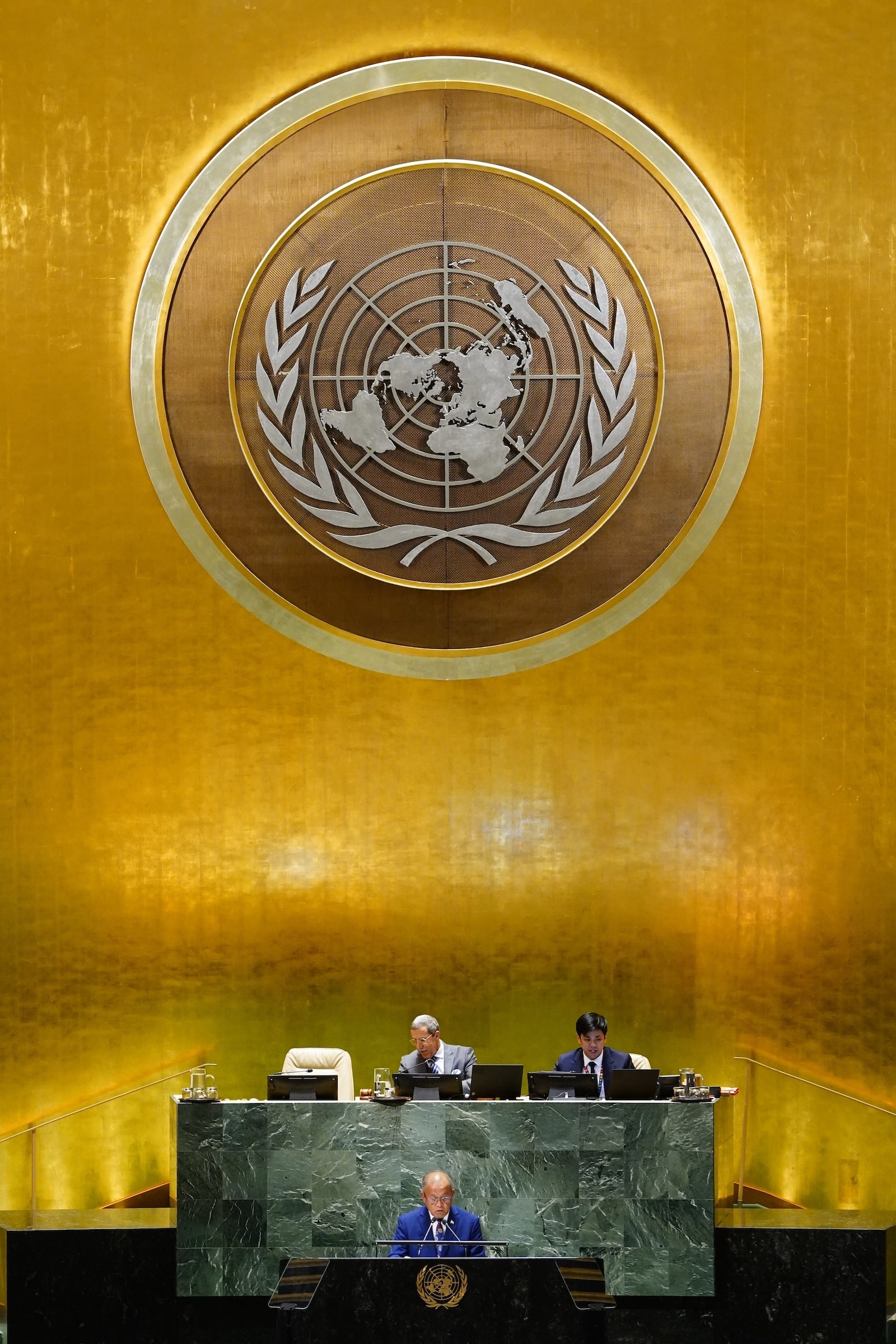 A large UN seal in a gold room under which a man in a suit speaks at a podium