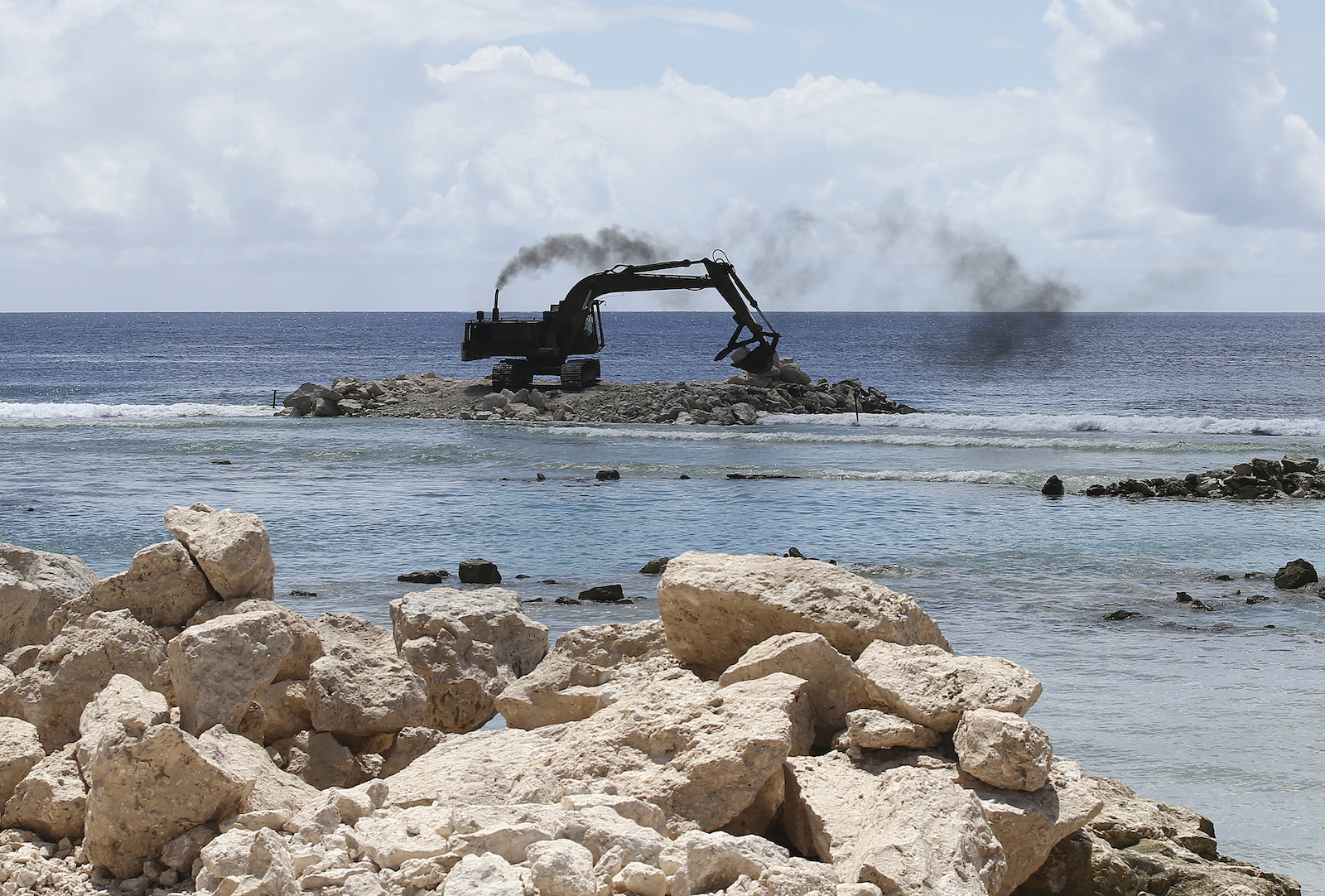 An exxcavator sits on a shallow part of the ocean with rocks in the foreground