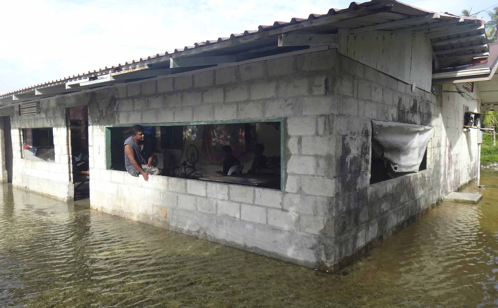 A man sits in the window of a cinderblock home with flood waters all around