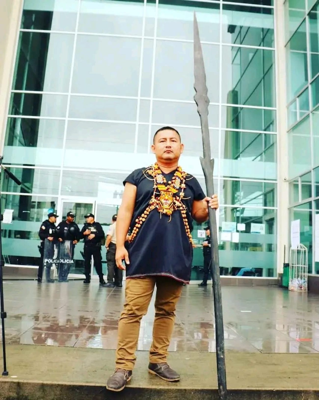 a man stands in front of a glass building holding a long carved stick.