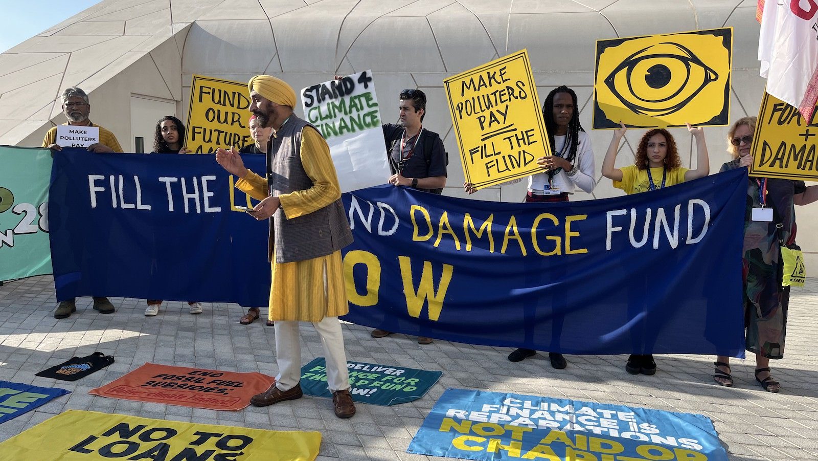 A man in a yellow turban and shirt stands near protesters and banners with messages related to climate reparations