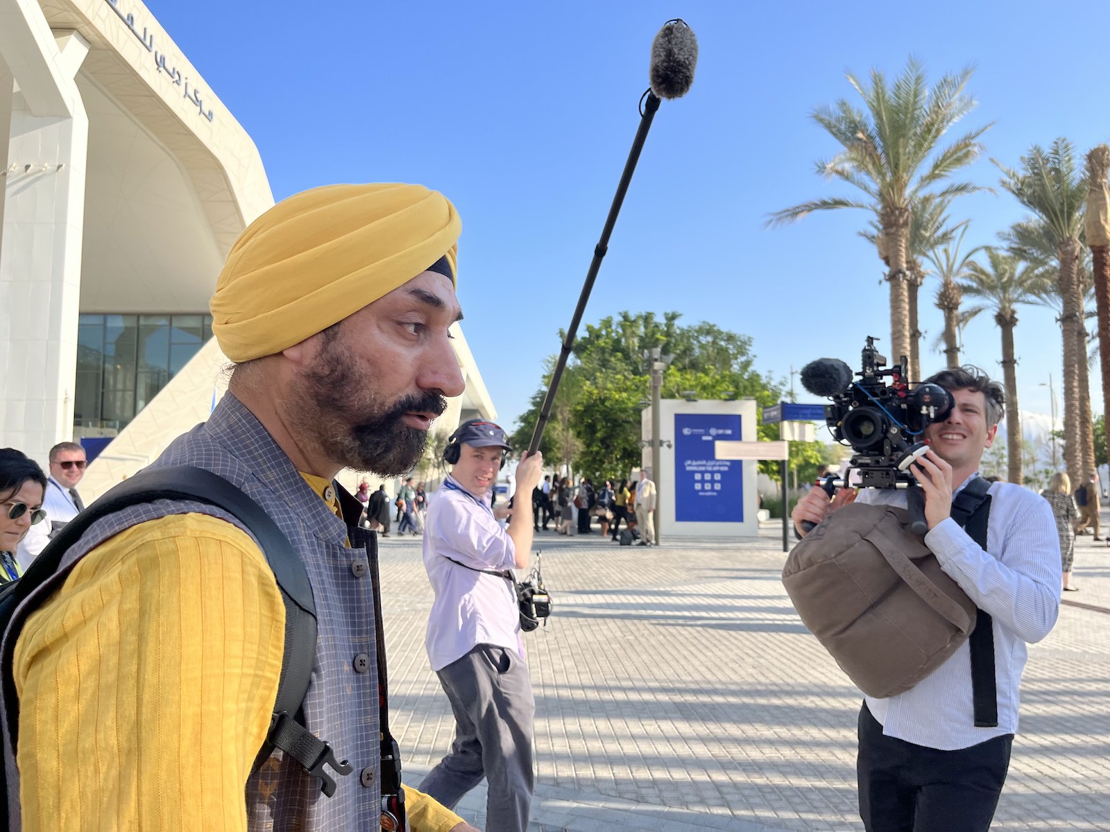 A man in a yellow turban and yellow shirt walks under a boom mic while another person films him