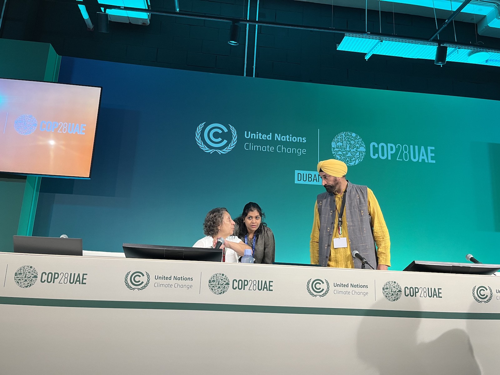 A man in a yellow turban and shirt stands behind a long table marked with COP28UAE. He is looking at two women also behind the table.