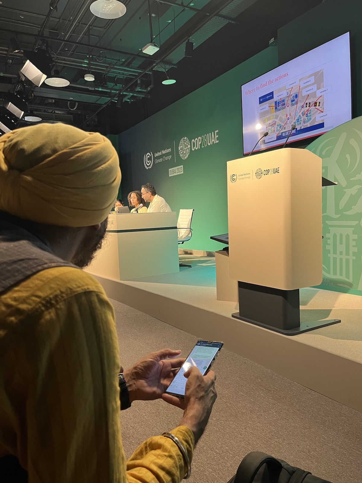 A man in a yellow turban and shirt holds a cell phone while sitting in the front row of a conference session at COP28UAE