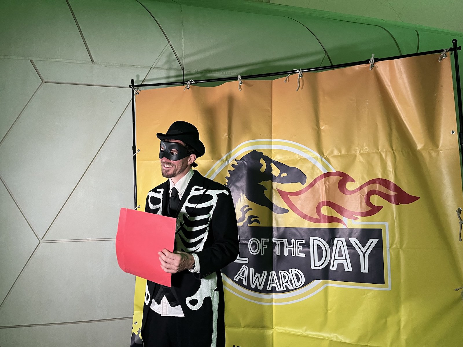 A man wears a bowler hat, black half-mask, and skeleton bone coat while holding a red sign and standing in front of a backdrop with a tyrannosaurus rex breathing fire.