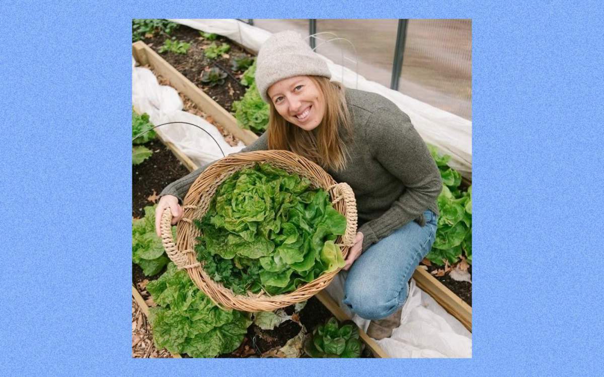 A blond woman in a hat and sweater crouches in between garden rows, smiling and holding up a basket of fresh green lettuce.